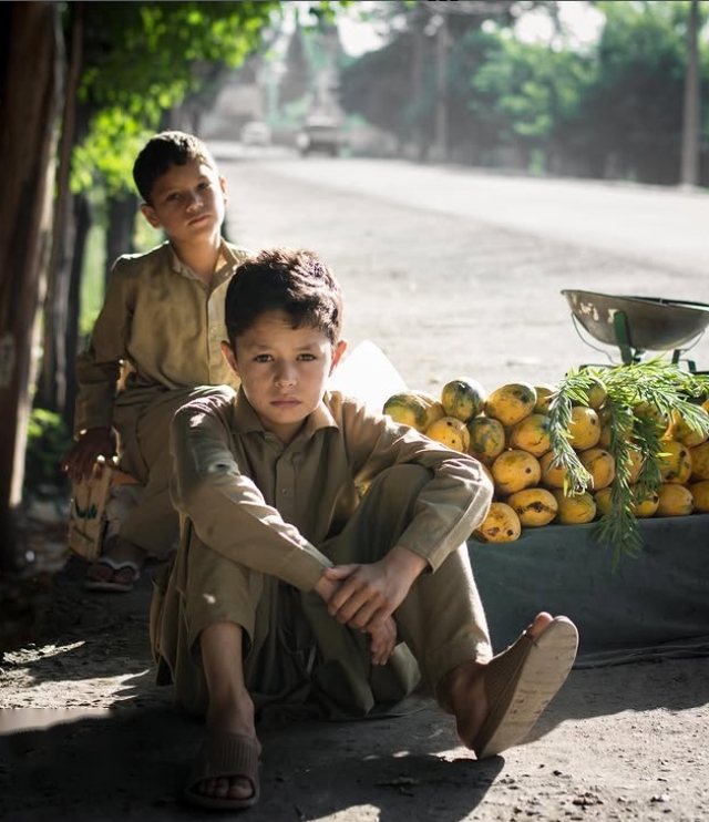 Two boys resting beside a roadside stall stacked with mangoes in warm afternoon light