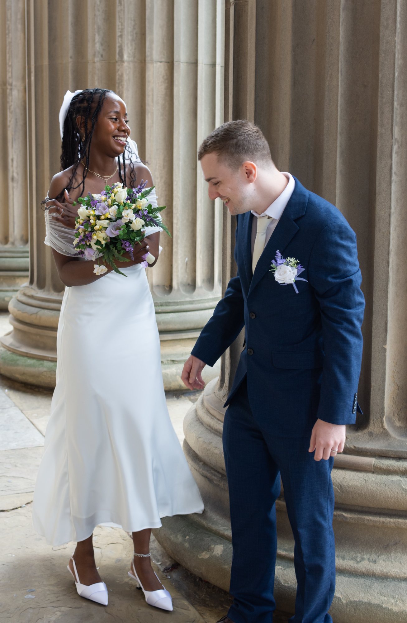 A couple laughing together beside tall stone columns, the woman holding a pastel bouquet